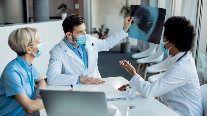 Group of doctors cooperating while analyzing X-ray of a patient at the hospital during coronavirus pandemic. Focus is on male doctor.
